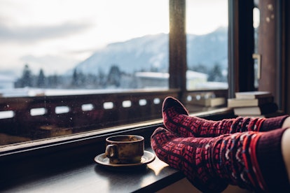 Feet in woollen socks by the Alps mountains view. Woman relaxes by mountain view with a cup of hot d...