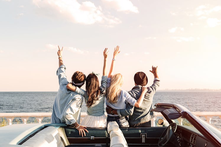 A group of friends stand next to their convertible with their arms up looking at the ocean on a holi...