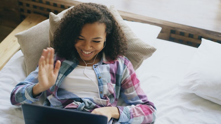 Cheerful mixed race girl having video chat with friends using laptop camera while lying on bed