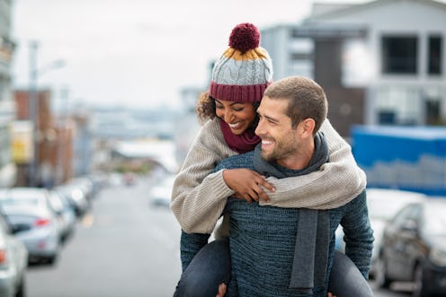 Smiling man giving piggyback ride to woman in the city. Young multiethnic couple in cold clothes wal...