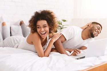 Young millennial african-american couple laughing, getting crazy lying on bed at home and using lapt...