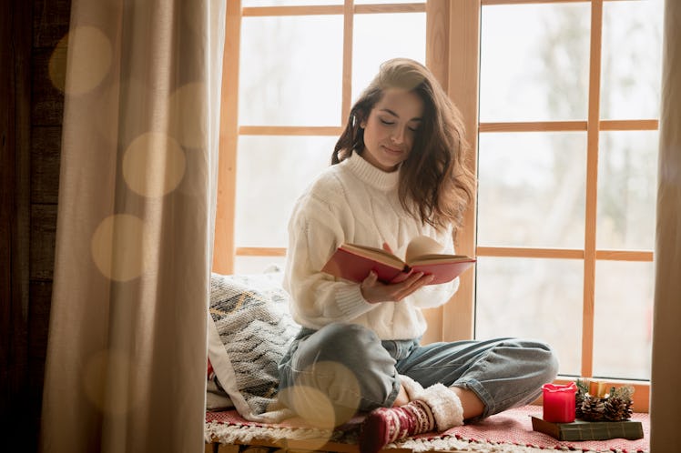 brunette woman in warm sweater and socks reading book sitting on windowsill in room decorated for ce...
