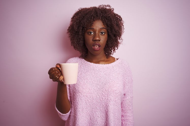Young african afro woman holding cup of coffee standing over isolated pink background scared in shoc...