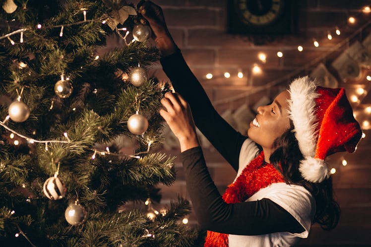 A woman wearing a Santa hat smiles while she decorates her Christmas tree.