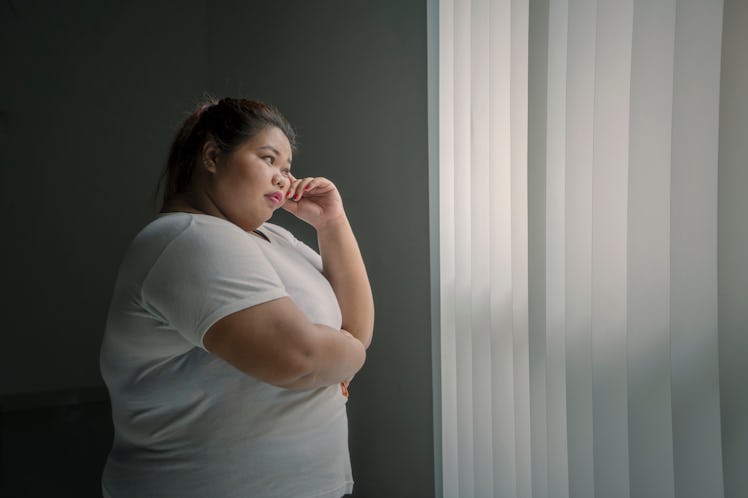 Picture of pensive obese woman looking out the window while standing in the dark room