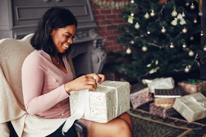 A woman in a fancy pink dress opens a gift while sitting near a Christmas tree.