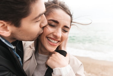 Happy lovely young couple wearing coats walking at the beach, holding hands, embracing, kissing