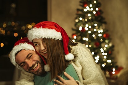 A couple wearing Santa hats hug each other in front of a Christmas tree during the holidays.