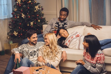 A group of friends in ugly Christmas sweaters relax in the living room next to a Christmas tree.