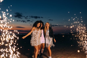 Three friends in sparkly dresses dance on the beach on New Year's Eve around sparklers and fireworks...