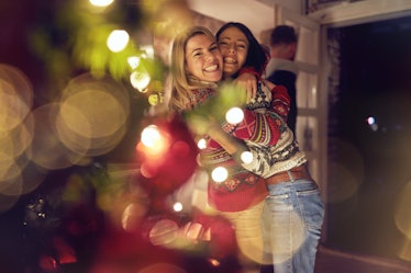 Two girls wearing ugly Christmas sweaters smile and hug next to a Christmas tree, for which they nee...