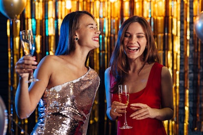 Two girls laugh on New Year's Eve in front of a shiny gold backdrop holding their champagne flutes.