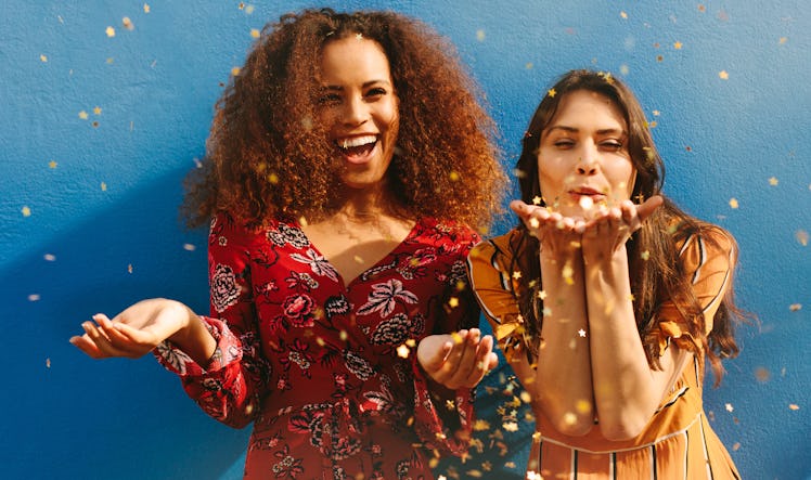 Two girls laugh and blow glitter and confetti into the air.