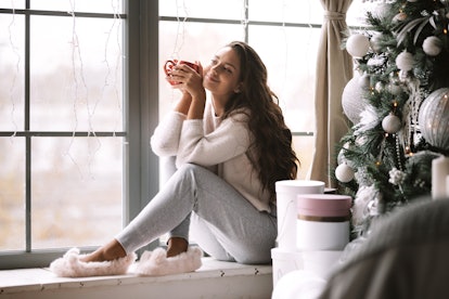 A woman dressed in comfy clothes and fuzzy slippers sits on a windowsill and holds a red mug at a be...