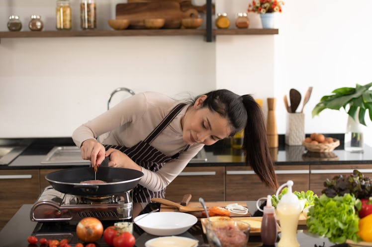 A brunette woman wearing a turtleneck and apron cooks a meal in her kitchen.