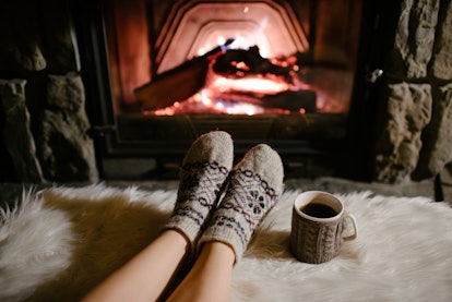 A woman in wool socks relaxes her feet next to a fireplace, blanket, and cup of hot tea in a cabin. 