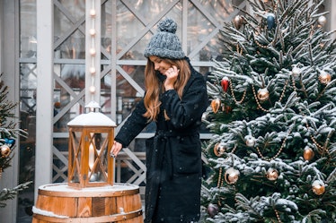A woman with long, wavy hair and a black jacket poses next to a snowy Christmas tree and lantern in ...