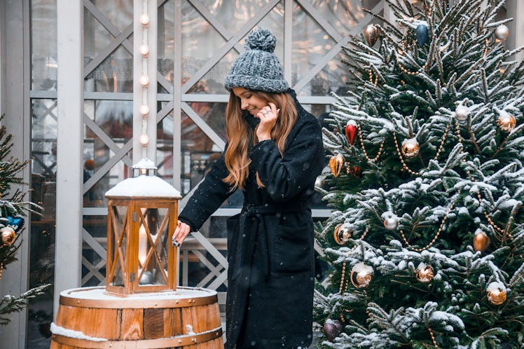 A woman with long, wavy hair and a black jacket poses next to a snowy Christmas tree and lantern in ...