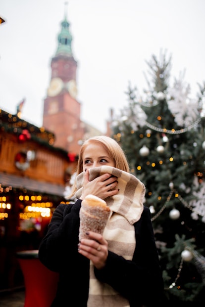 A blonde woman holds a sugary treat in a holiday market while covering her face with her scarf in a ...
