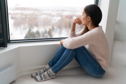 Depressed young girl feeling sad an lonely, anxious looking out the window in winter. Unhappy Asian ...