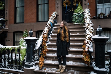 A brunette woman looks up and stands on snowy steps of a NYC apartment in the winter.