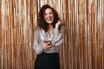 Positive girl in silver outfit holding phone and laughing