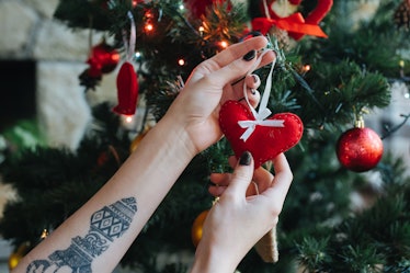 A woman with tattoos hangs a heart-shaped ornament on a Christmas tree.