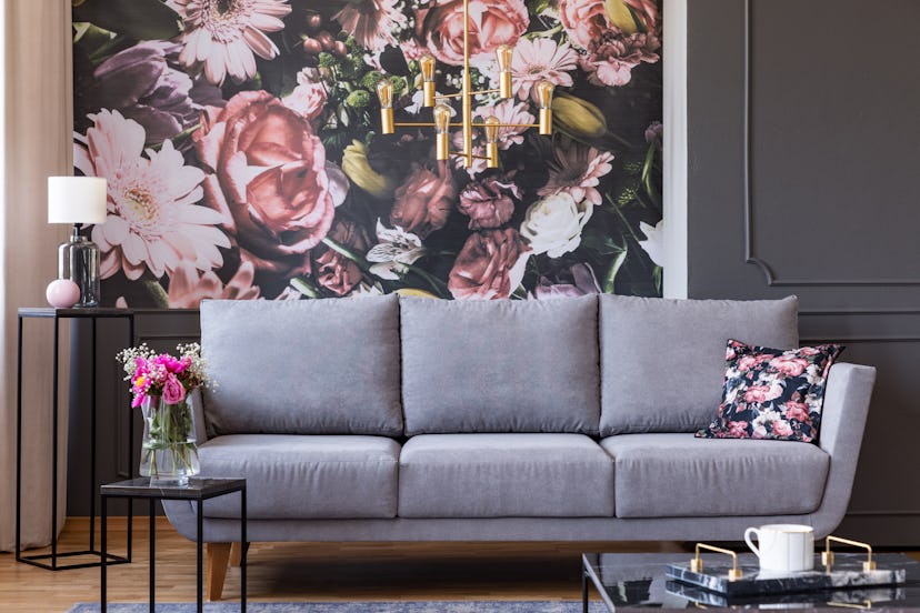 Flowers on black table and grey sofa in living room interior with lamp and wallpaper. Real photo