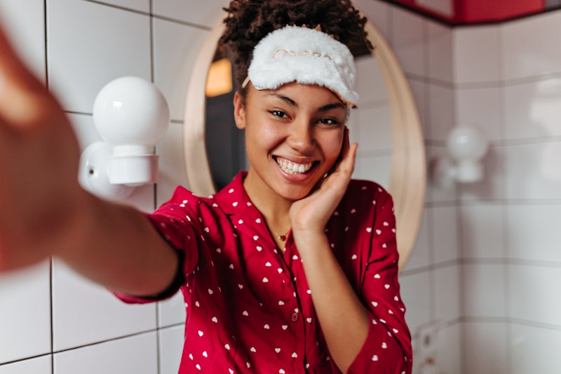 Charming dark-skinned woman in sleep mask is smiling, looking at camera and taking selfie