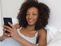 Young cheerful comfortable woman texing message on smartphone in the bed at home. Afro hair black be...
