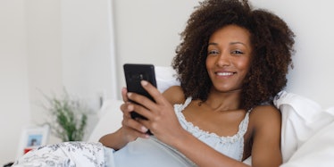 Young cheerful comfortable woman texing message on smartphone in the bed at home. Afro hair black be...