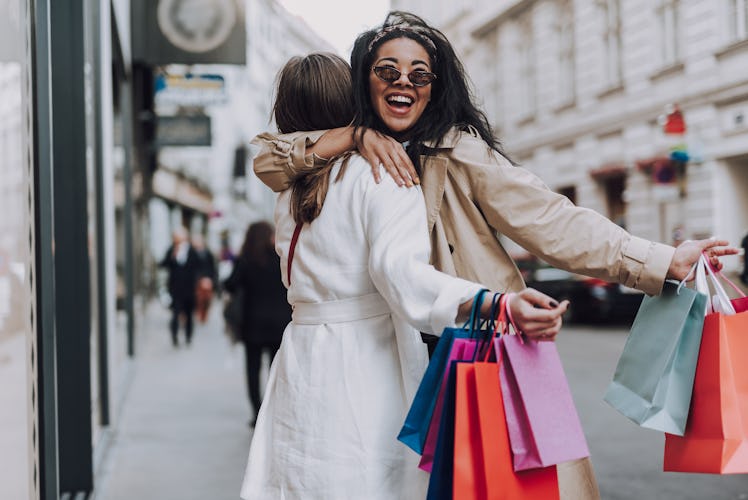 Two girls hug in the middle of a city with colorful shopping bags in their hands.