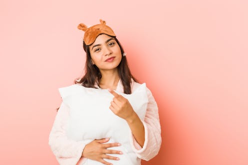 Young indian woman wearing a pajamas and sleep mask isolated holding a pillow.