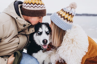 A stylish couple kisses their husky while hanging out by a lake in the middle of winter.