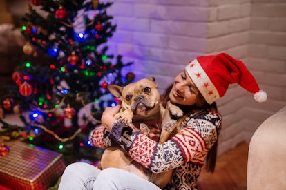 A woman wearing a Santa hat and holiday sweater smiles and holds her dog next to Christmas presents ...