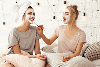 Two girls do facials while hanging out in bed and smiling.