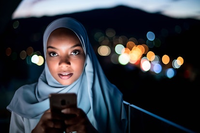 Young modern Muslim woman wearing scarf veil on urban city  street at night texting on smartphone wi...