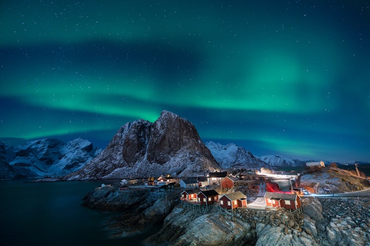 Lofoten, Norway at night features the Northern Lights, snowy mountains, and tiny red cabins lit up. 