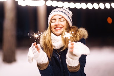 Cheerful young woman holding a sparkler in hand  in the winter forest. Happy cute girl in knitted ha...