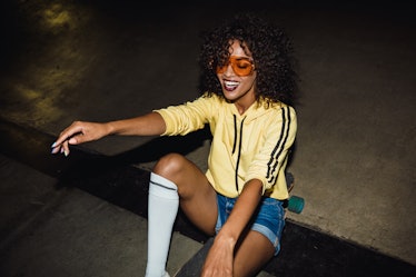 A woman in orange sunglasses and street clothes sits on a skateboard and smiles at night.
