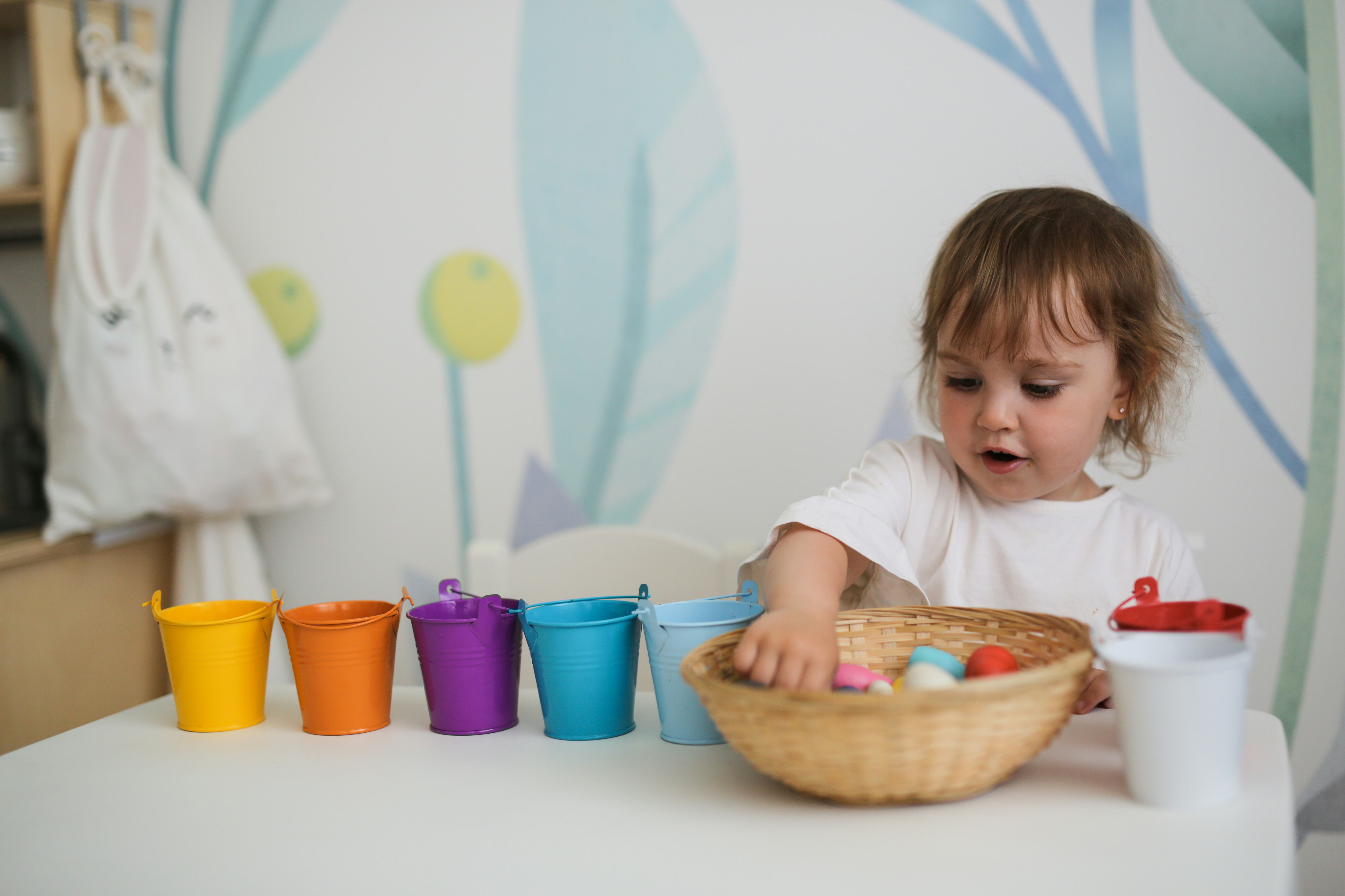 charming toddler girl sorts colorful toys by colored buckets at the table