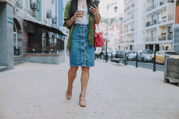A woman in casual work attire walks down a city street with coffee in her hand.