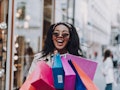 A smiling woman in sunglasses holds a bunch of colorful shopping bags on a sunny day.