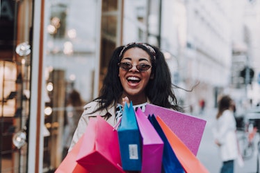 A smiling woman in sunglasses holds a bunch of colorful shopping bags on a sunny day.