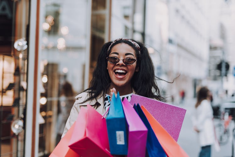 A smiling woman in sunglasses holds a bunch of colorful shopping bags on a sunny day.