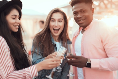 A group of three friends smile and toast their champagne flutes outside on a sunny day.