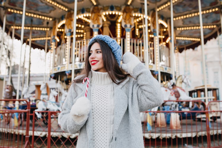 A stylish woman in a grey jacket and blue knitted hat holds a candy cane and poses in front of a car...