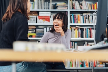 Two women best friends laugh while sitting at a table at work in front of a bookshelf.