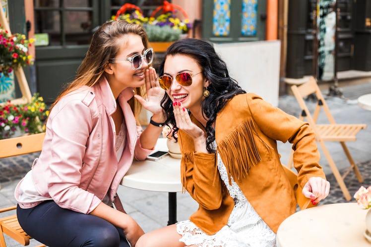 Two best friends whisper secrets to each other while sitting at an outdoor cafe.