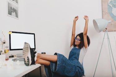 A woman in denim overalls sits at her desk and stretches her arms above her head.